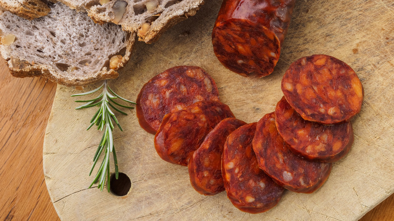 Sliced chorizo on wooden cutting board next to bread and sprig of rosemary.