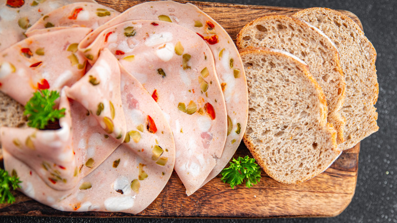 Thinly sliced mortadella with bits of pepper and pistachios next to bread on cutting board.