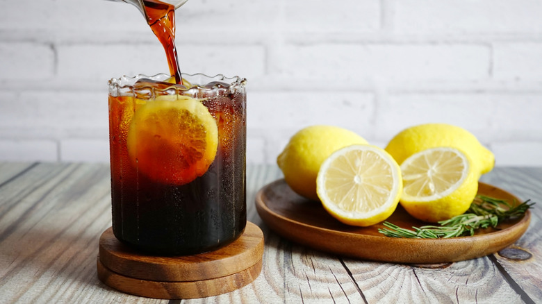 A glass of sparkling sweet tea being poured on a wooden table alongside three lemons