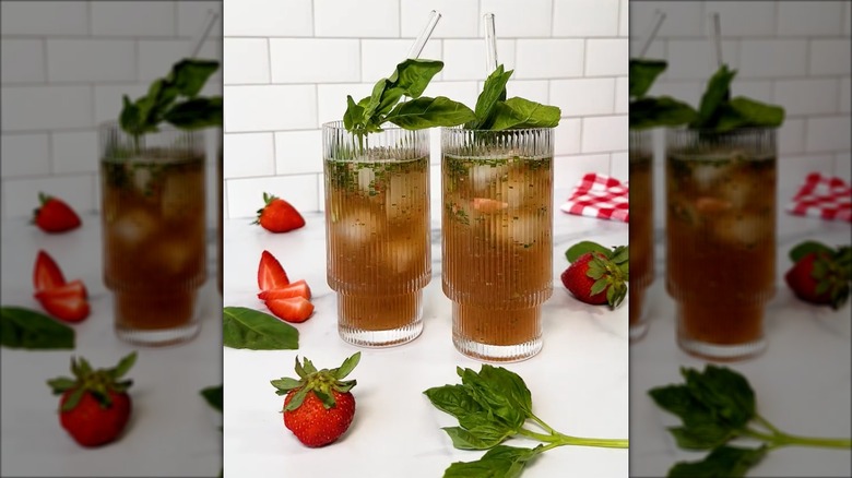 Two glasses of Strawberry Basil Sweet tea surrounded by strawberries and basil on a white counter and white tile background