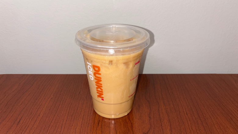 A light brown iced coffee in a clear plastic Dunkin' cup has condensation and drips down the cup