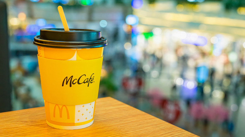 McCafe takeaway cup with wooden stick placed on table, blurred urban environment in the background.