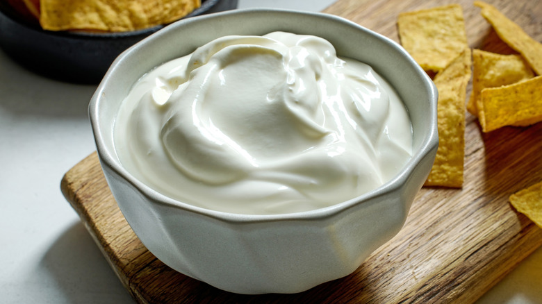 A dish of sour cream on a wooden cutting board with tortilla chips in the background