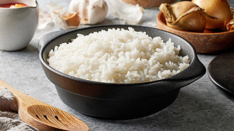 White rice in a black bowl with a wooden spoon on the table and ingredients in the background