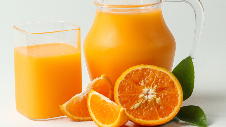 An orange cut in half with two slices sits next to green leaves and glass and pitcher full of orange juice on a white background