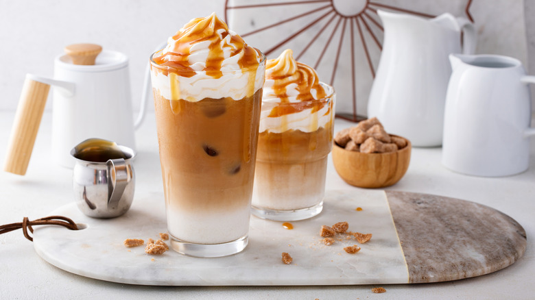 Two fancy coffee drinks in clear glasses topped with whipped cream and caramel sauce surrounded by coffee jugs and sugar