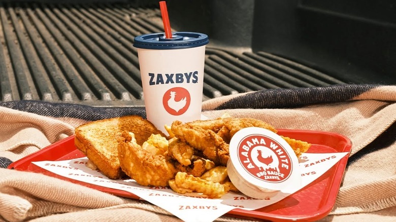 An orange Zaxby's tray on a striped blanket with food, a drink, and a container of Alabama white barbecue sauce