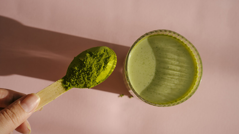 A person with a spoonful of matcha powder and a glass of matcha on a pink background