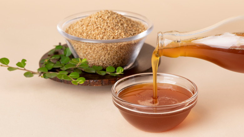 Sesame oil poured into a glass bowl with a glass bowl of sesame seeds in the background
