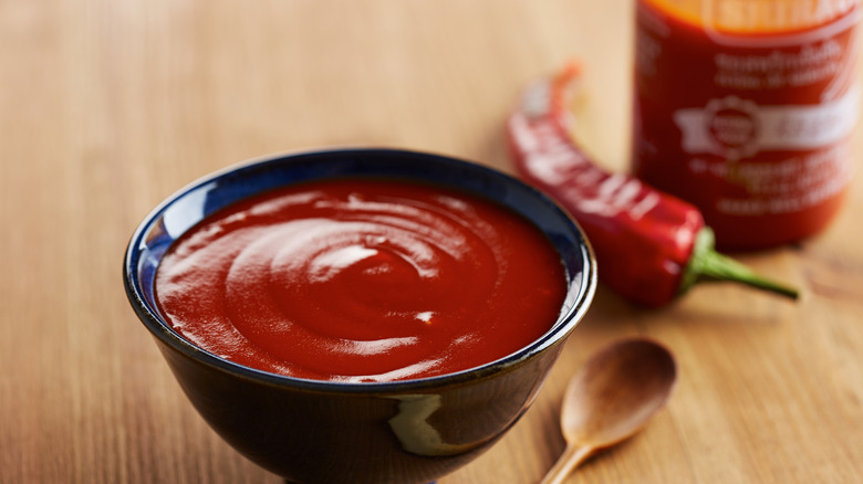 Small bowl of Sriracha sauce with a pepper and bottle in background.