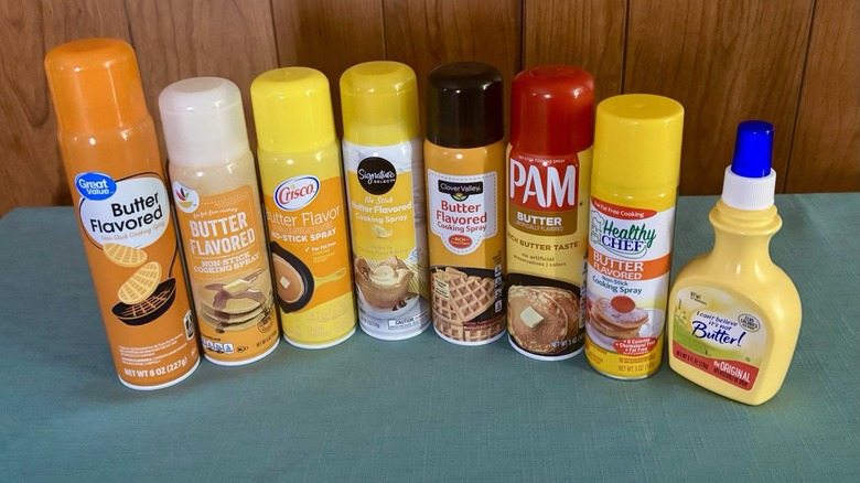 Eight butter flavored non-stick sprays in a line-up. From left - Great Value, Stop & Shop, Crisco, Signature Select, Clover Valley, PAM, Healthy Chef, and I Can't Believe It's Not Butter!