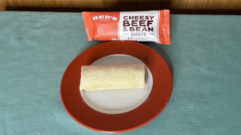 Red's Cheesy Beef & Bean Burrito on a red plate, in front of the burrito's packaging