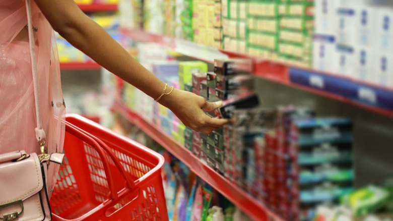 A woman reaching for an item on a supermarket shelf carrying a red basket