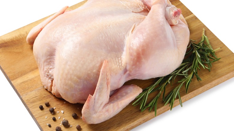 A whole raw chicken on a cutting board with fresh rosemary and peppercorns