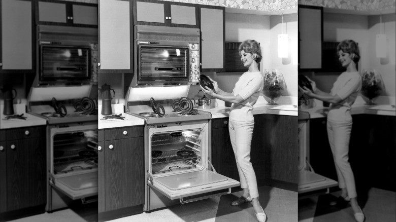 Black and white photo of a woman looking at a clean dish while standing by a range with a top oven