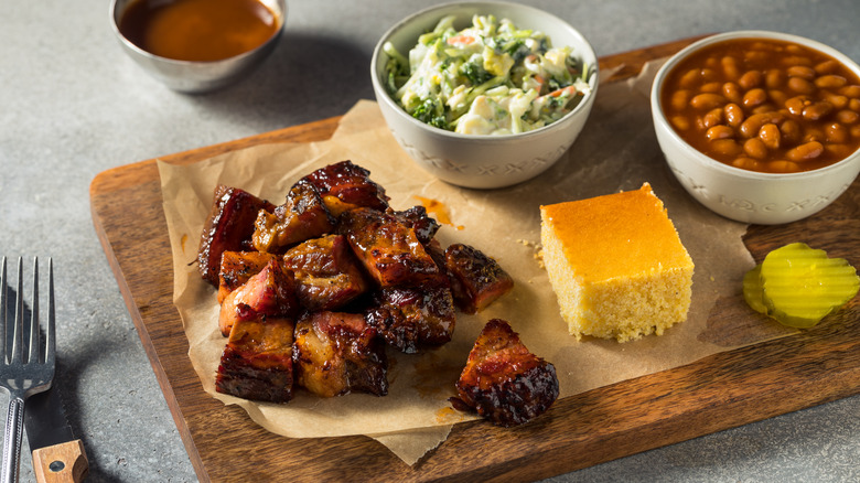 Brisket burnt ends with salad, beans, cornbread, and pickle slices on a wooden board