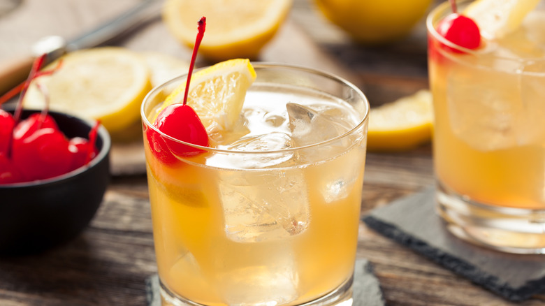 A pale yellow cocktail in a rocks glass with ice, a lemon wedge and cherry.