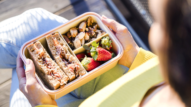 Person holding a bento-style lunch box with nuts, berries, and a sandwich