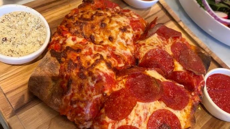 A square pepperoni pizza on a wooden board with bowls of sauce and cheese on side