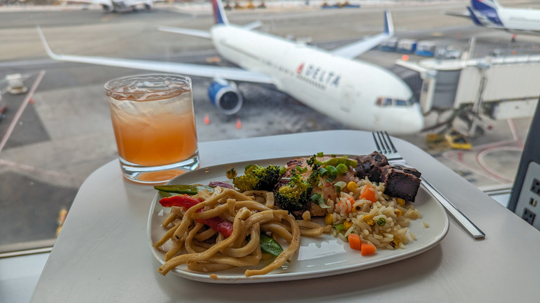 A plate of noodles and rice with a cocktail on a table in front of a window with a view of a plane parked at a gate at airport