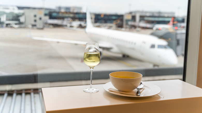 A cup of soup and a small glass of wine on a table in front of a window with a view of a plane outside parked at a gate
