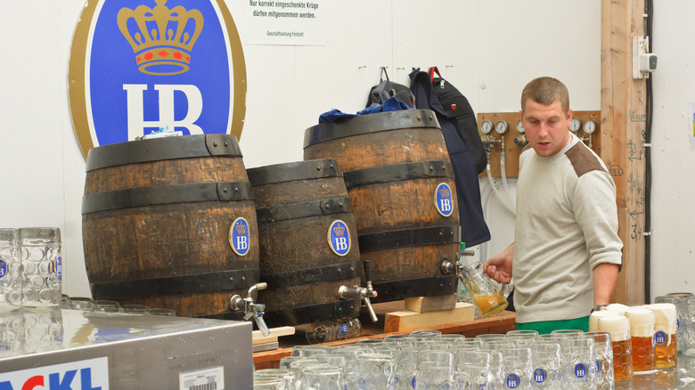 A man fills stein glasses with Oktoberfestbier at a beer hall featuring the Hofbräu logo appearing on the wall, the wooden barrels, and the glasses