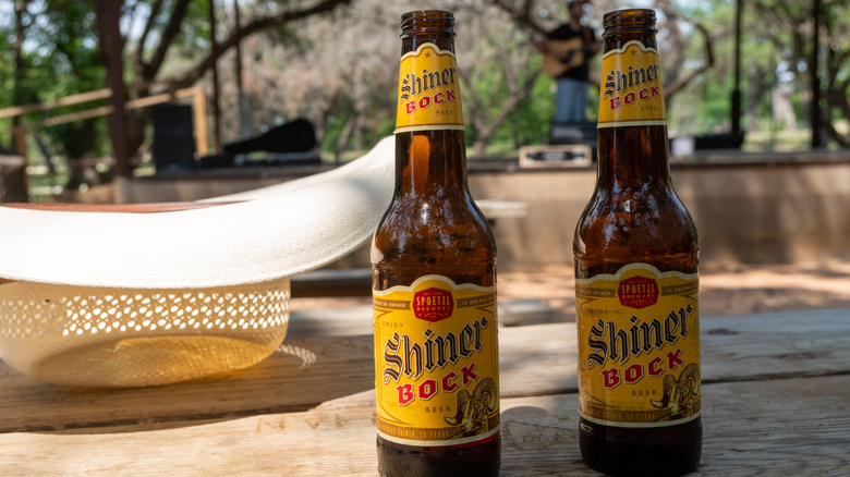 Two bottles of Shiner Bock rest on a table, with a picnic basket and trees in the background