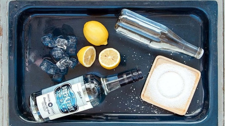 A bottle of Deep Eddy on a tray with melting ice, a cut lemon, a bottle of soda water, and tray of salt