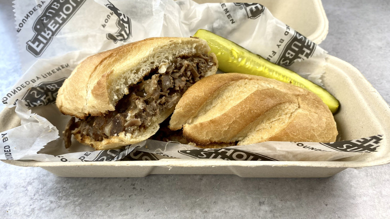 Firehouse Subs Steak & Cheese sandwich cut in half in the take-out container next to a pickle spear.