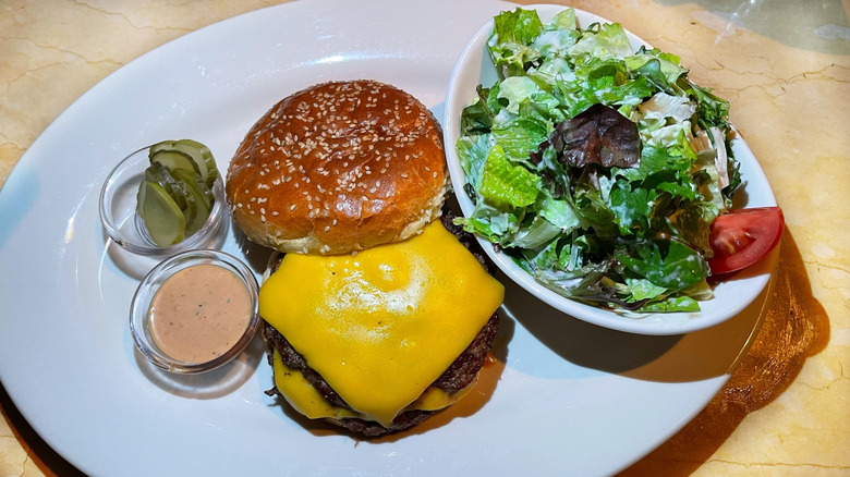 White plate consisting of side salad with a bright red tomato and a cheeseburger with the top bun slid off