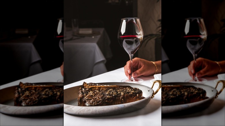 Porterhouse steak on a metal platter and a person holding a glass of red wine