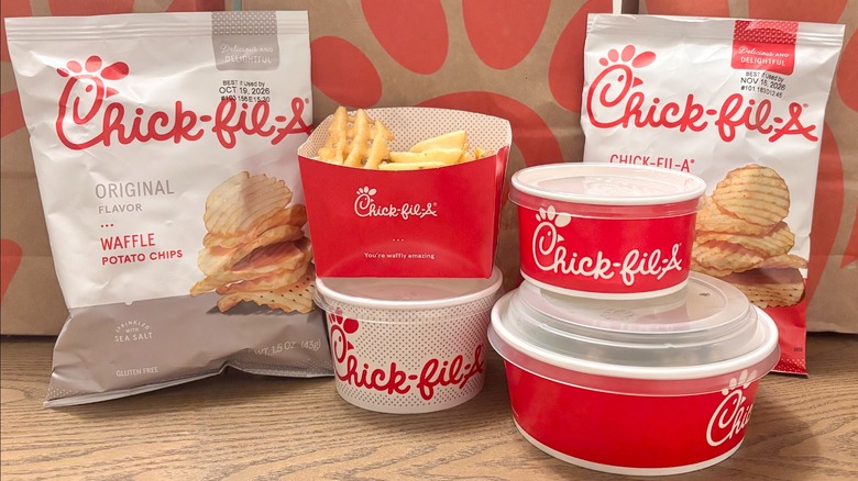 A variety of Chick-fil-A food containers, fries, and two bags of Chick-fil-A chips in front of brown paper Chick-fil-A takeout bags