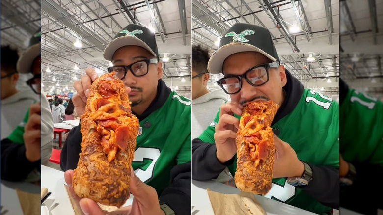 Man shows off and takes a bite of a Costco hot dog stuffed in a chicken bake topped with a pizza slice