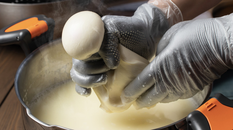 Two gloved hands pulling fresh mozzarella curds in a warm brine in a pot