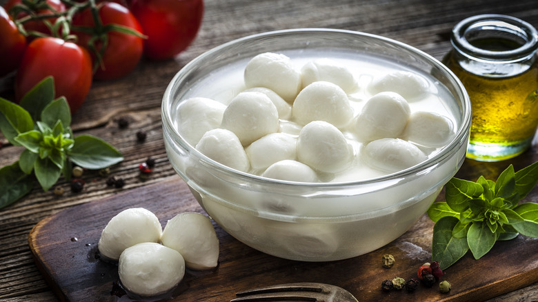 A bowl filled with fresh small mozzarella balls on a wooden table next to oil, tomatoes, and fresh basil