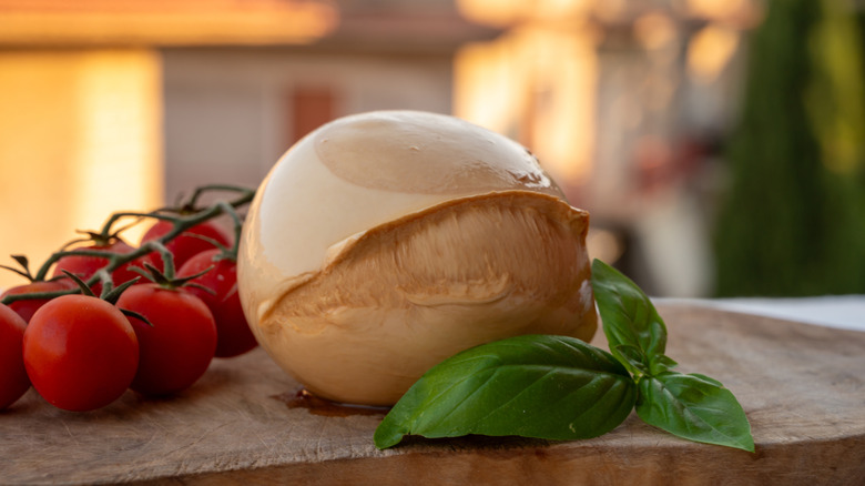 A whole ball of smoked mozzarella on a wooden board next to fresh tomatoes and basil leaves