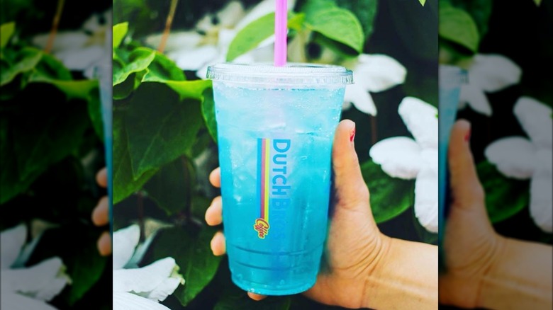 A person holding an Ocean water drink in front of a white flowering bush