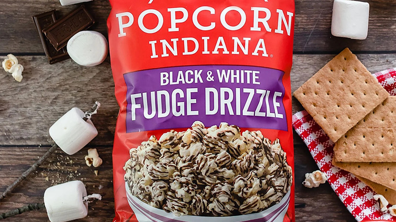 A close up of a bag of Popcorn Indiana Black & White Fudge Drizzle Popcorn on a table with graham crackers, marshmallows, and pieces of chocolate