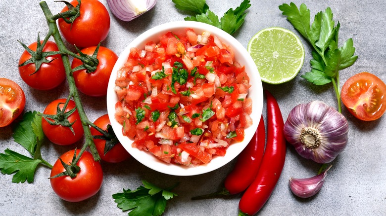 A bowl of pico de gallo surrounded by its essential ingredients, such as tomato, onion, cilantro, lime, and chile pepper