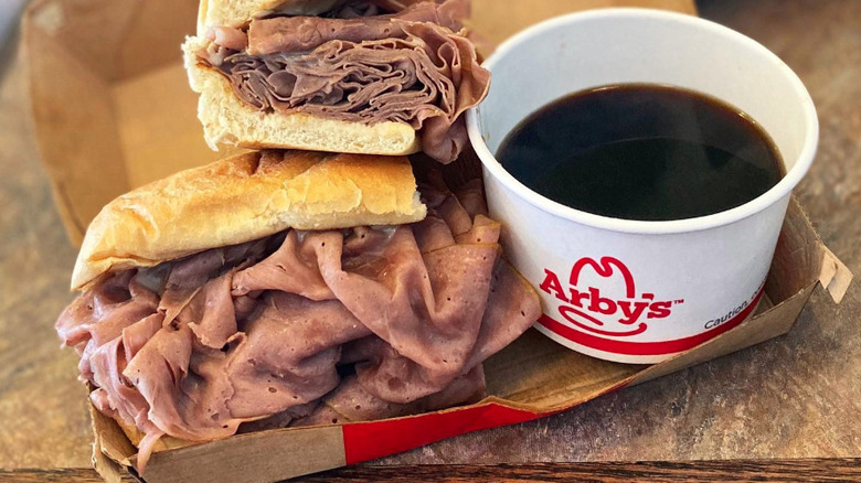 Arby's French Dip & Swiss sandwich stacked with roast beef, served in a cardboard tray with a cup of au jus dipping sauce
