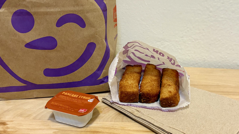 An order of French Toast Sticks from Jack in the Box sitting on some napkins next to a Jack in the Box bag and pancake syrup