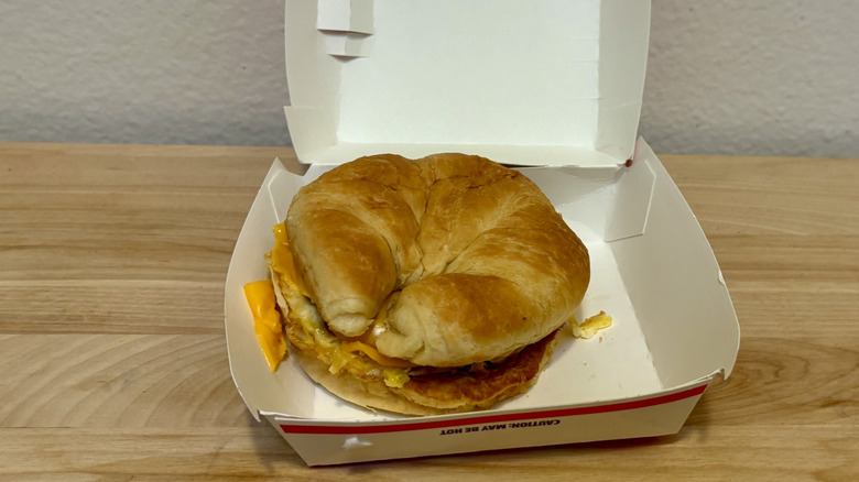A Sausage Croissant from Jack in the Box sitting on a wood table inside of its box