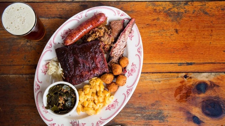 A plate of ribs, brisket, and sausage with sides alongside a beer on a wooden tabletop