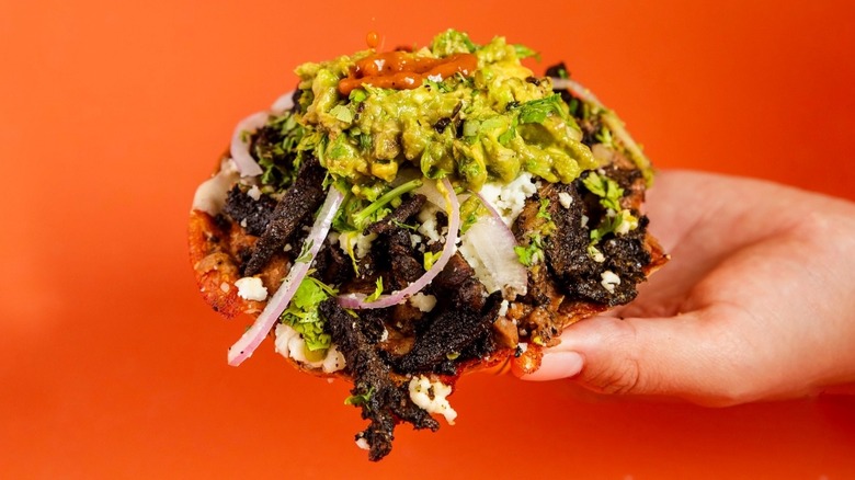 A hand holds a taco shell piled high with toppings on an orange background