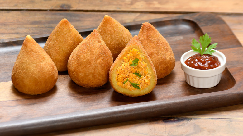 six teardrop shaped croquettes on a wooden serving tray sit next to a ramekin of ketchup with a sprig of fresh herb on top - one of the croquettes has been cut open to reveal it's cross-section of spicy chicken and herbs