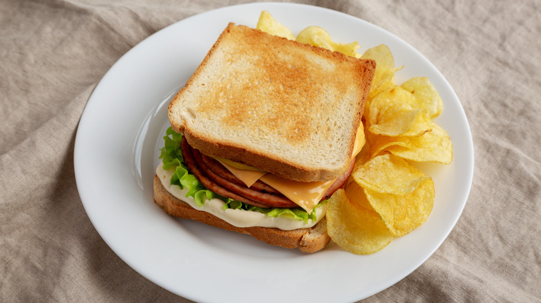 A toasted fried bologna sandwich with mayonnaise, cheese, and lettuce on a plate, paired with potato chips