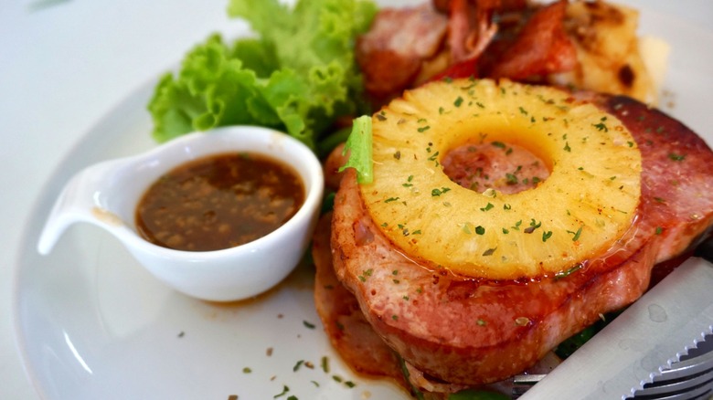 A slice of ham steak with grilled pineapple on top and a small sauce boat beside it - lettuce and mash in the background and a knife and fork in the foreground