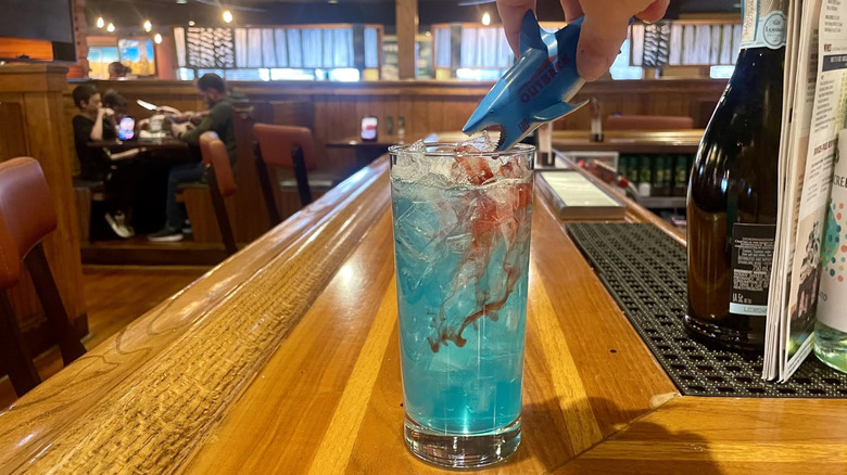 Hand pouring grenadine into Outback Steakhouse's Aussie JAWSsie cocktail from top shark's mouth