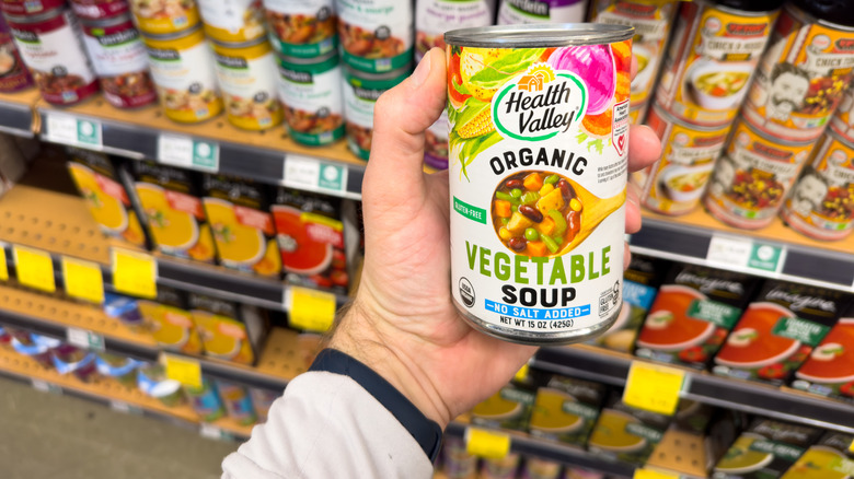 A person holding a can of soup in front of the Whole Foods market soup aisle