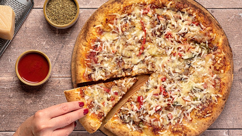 hands removing a pizza slice from a pizza topped with bbq pork, peppers, and cheese. pizza is sitting on a counter next to oregano and various sauces